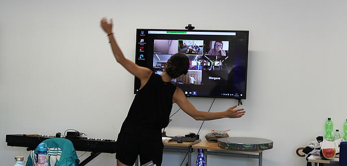 Jugendlicher mit seitlich erhobenen Armen vor einem Bildschirm, der eine Videokonferenz zeigt.