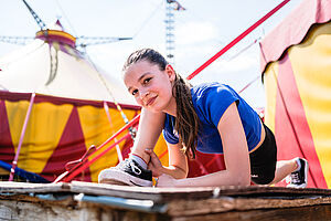 Titelbild des BKJ-Förderprogramms „Start2Act“ Ein Kind macht Aufwärmübungen vor einem Zirkuszelt. Es schaut direkt in die Kamera.