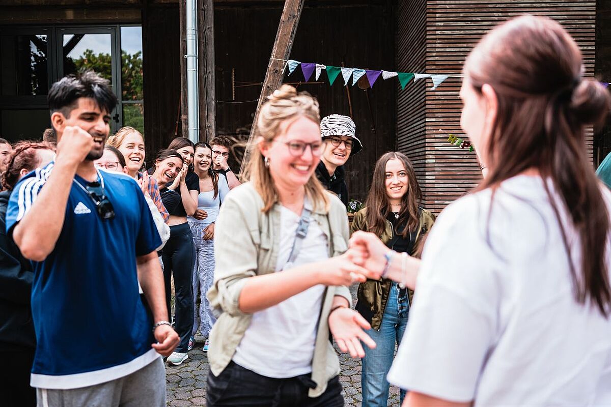 Mehrere junge Menschen, im Vordergrung zwei lachende junge Menschen, die Stein Schere Papier spielen