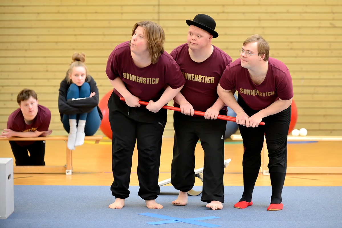 Drei Artist*innen des Circus Sonnenstich stehen nebeneinander, beugen sich leicht nach vorn und halten gemeinsam einen roten Stab quer vor sich.