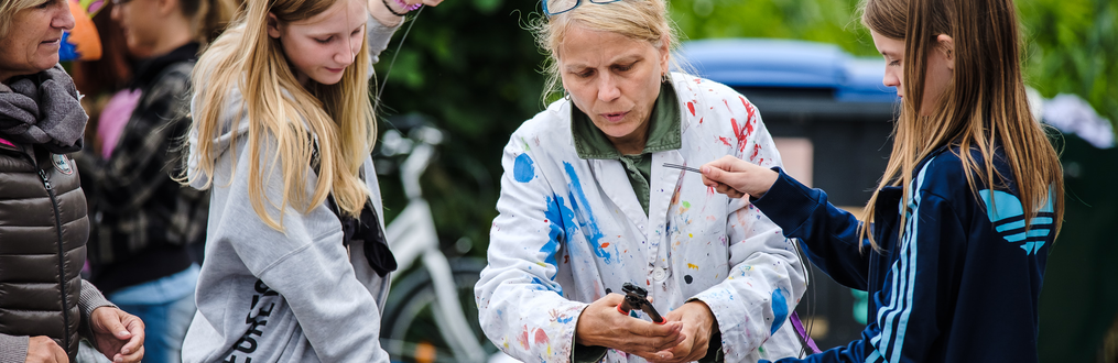 Fachstelle Kulturelle Bildung Zwei Erwachsene und zwei Jugendliche, bekleidet mit Schutzkittel voller bunter Farbflecke, werkeln draußen gemeinsam an einer Skulptur.